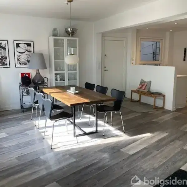 Spisebord med sorte stole står i et lyst rum med trægulv. Ved siden af står en reol og lamper. Væggen er dekoreret med sort-hvide fotografier. Ingen tekst er synlig.