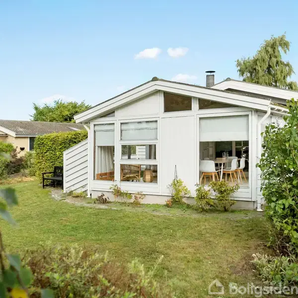 Hvidt hus står stille i en frodig have med grønne buske og en træramme. Indenfor ses et spisebord med stole. Sollys skinner gennem store vinduer.