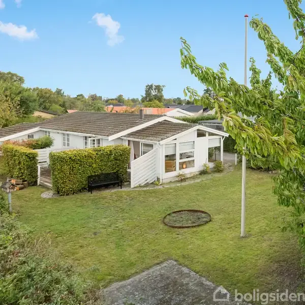 Huset står stille i en velplejet have med græsplæne og træer omkring. En flagstang er placeret ved siden af huset. Det er en lys dag med få skyer på himlen.