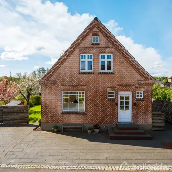 Murstenshus med trekantet gavl har tre vinduer og en hvid dør foran. Det står på en brolagt plads med blomstrende træer og grøn beplantning i baggrunden under en blå himmel. Ingen tekst.