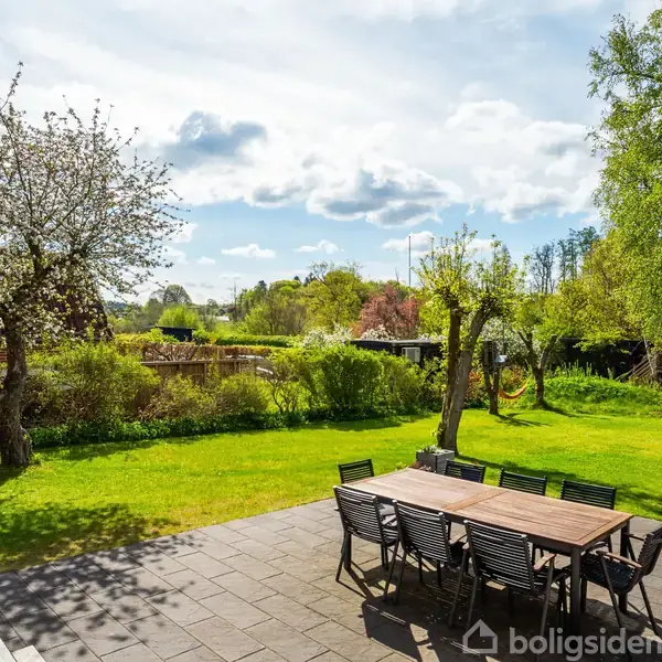 Et træbord med stole står på en flisebelagt terrasse. Omgivet af en frodig have med grønne træer og buske under en skyet blå himmel.