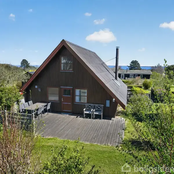 Hus af træ står stille i en grøn have med terrasse og havemøbler. Omkring er der buske og træer under en blå himmel med skyer.
