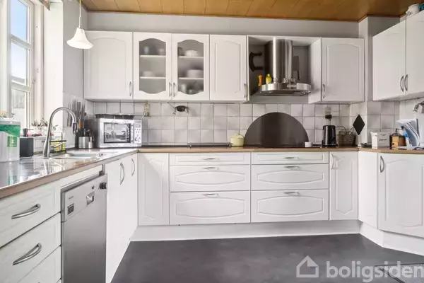 Et hvidt køkken med skabe, en opvaskemaskine og køkkenvask. Der er en mikrobølgeovn og køkkenredskaber på bordet. Store vinduer slipper dagslys ind, hvilket skaber en lys atmosfære.