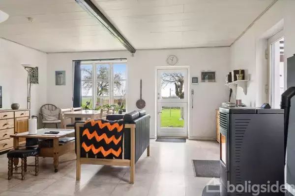 Stue med sofa med orange tæppe, træbord med stole, og en brændeovn. Store vinduer og glasdør åbner ud mod en have med græs og træer. Minimalistisk indretning.