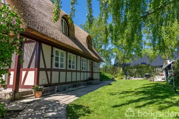 Et stråtækt bindingsværkshus står stille i en solrig have med grøn græsplæne og træer. En terrasse med fliser ses ved siden af huset.