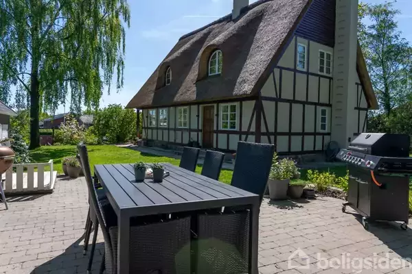 Et træbord med stole står på en solrig terrasse ved siden af en stor grill. I baggrunden er der et bindingsværkshus med stråtag omgivet af grønne planter.