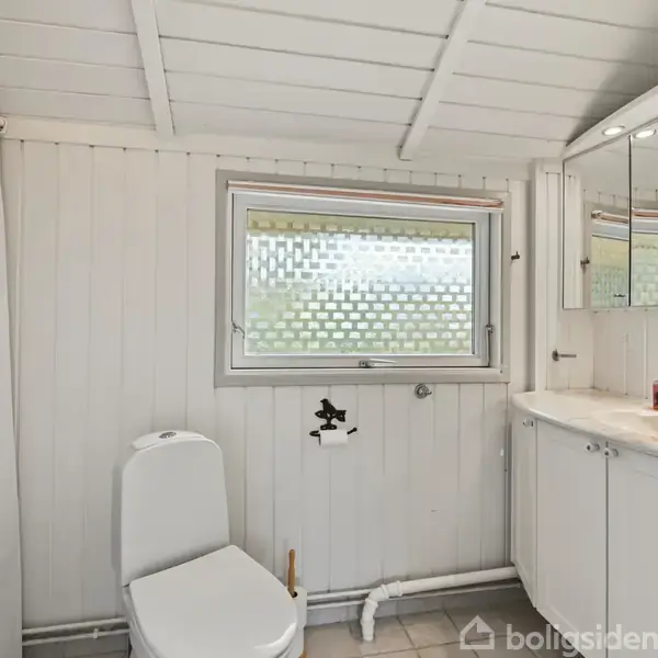Toilet står i et lyst badeværelse med hvide vægge og fliser. Til højre er en stor vask med spejlskab. Et vindue med mønstret glas lader lys komme ind.