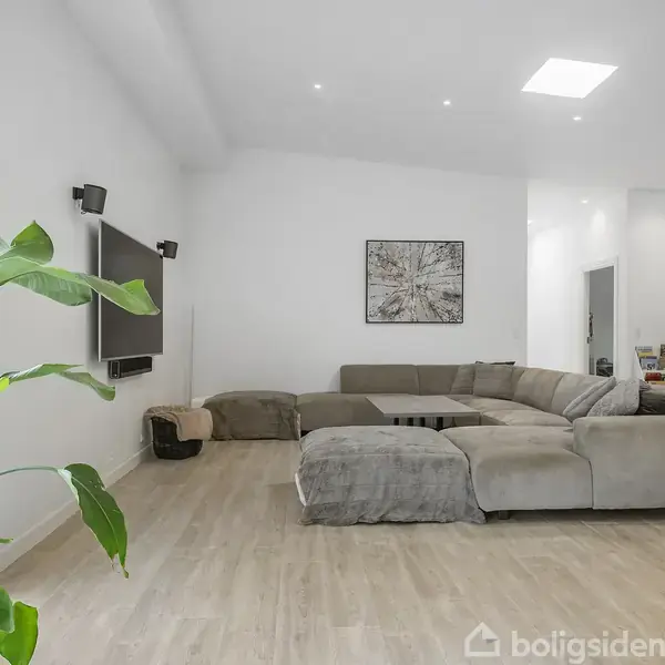 En stor, grå sofa står i en lys stue med trælaminatgulv og en grøn plante ved vinduet. Et TV hænger på væggen, og et kunstbillede er centralt placeret.
