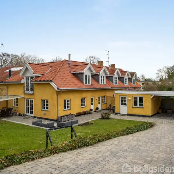Gulmalet hus med rødt tegltag, fem kvistvinduer, en carport med bil og flisebelagt indkørsel. Beliggenhed i en frodig have med grønne planter og buske.