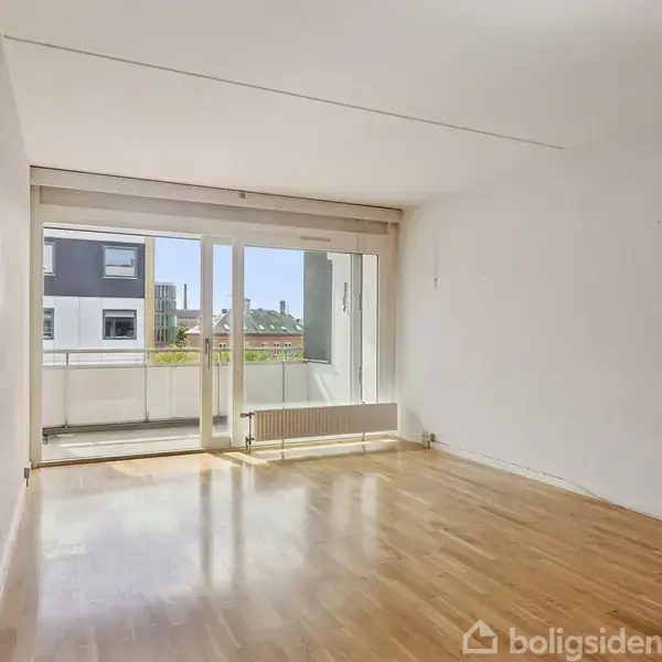 Stue med trægulv, lyst og tomt rum. Store glasdøre åbner ud til en balkon med udsigt til bygninger. Væggene er hvide, og der er lysindfald. En radiator findes under vinduet.