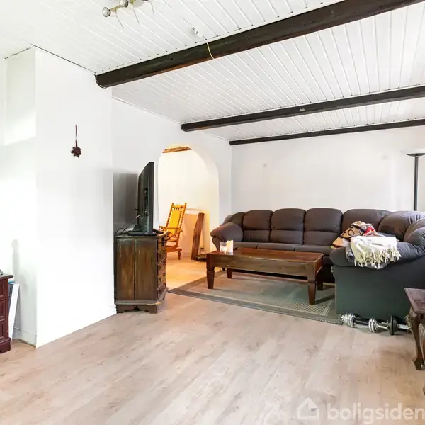 En stue med en mørk L-formet sofa, der står foran et sofabord. Ved siden af står en gulvlampe. I baggrunden ses en gyngestol og et fjernsyn. Væggene er hvide.