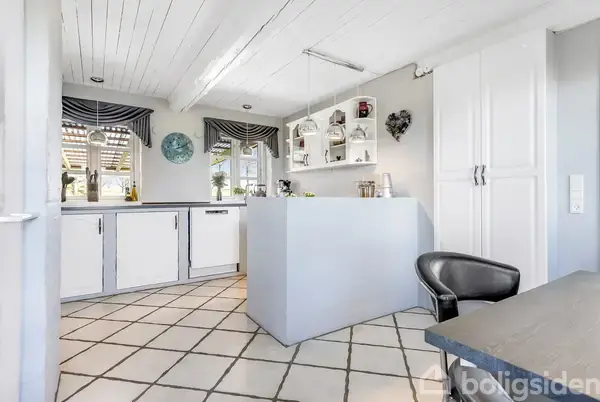 Køkken med hvidt interiør og flisebelagt gulv. Hvide køkkenskabe omgiver et centralt arbejdsområde. To vinduer med stribede gardiner giver udsigt til haven. Væggene har hjerteformet dekoration.