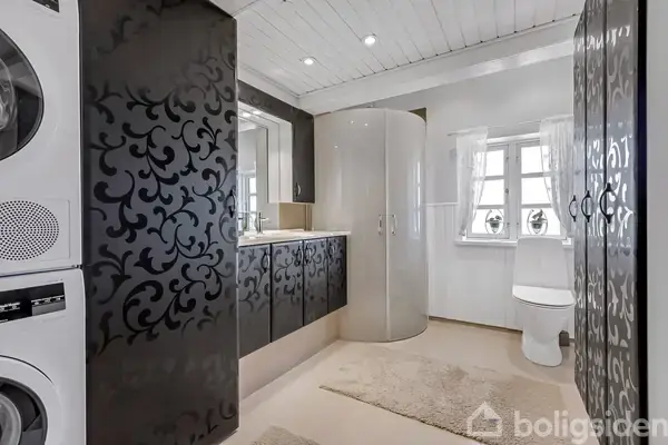 En elegant badeværelse med sort dekorerede skabsdøre, bruseniche, vaskemaskine og tørretumbler til venstre. Hvide vægge, træloft, tæppe på gulvet og toilet ved siden af vinduet med gardiner.