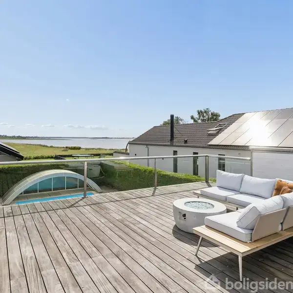 Terrasse med trædæk har en hjørnesofa og et lille rundt bord. Solpaneler ses på taget i baggrunden, og en pool kan skimtes bag en hæk med fjordudsigt i horisonten.