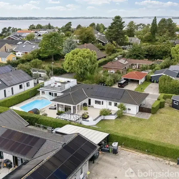 Huset har en swimmingpool i haven, omgivet af andre huse blandt frodige træer. Det ligger i en boligområde nær en sø med udsigt til skovklædte bakker i baggrunden.