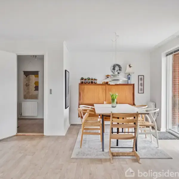 Et spisebord med seks stole står på et tæppe i et lyst rum. Der er en skænk med dekorationer i baggrunden og en åben dør til et andet rum. En klaveret står ved siden af døre til terrassen.