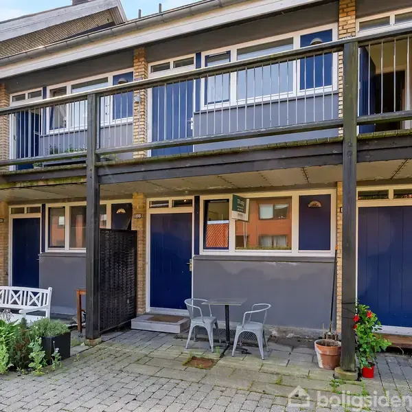 To-etagers rækkehus med blå døre, en balkon og vinduer øverst. Foran er et flisebelagt gårdhaveområde med hvid bænk, bord, stole og grønne planter.