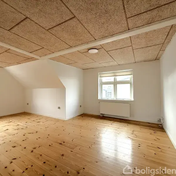 Et tomt værelse med trægulv og hvidmalede vægge, indeholder en radiator under et enkelt stort vindue, i en bolig med loft af træplader.