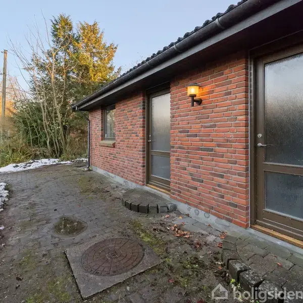 Rød murstensbygning med en tændt lanterne ved indgangen. En belagt sti med smeltende sne fører langs huset. Omgivet af en hæk og træer i en have.