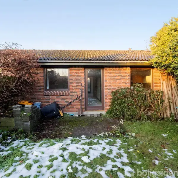 En rød murstensbygning med to vinduer. Foran er en have med snepletter, en trillebør og buske omkring. Det er lyst og ser ud til at være vinter.