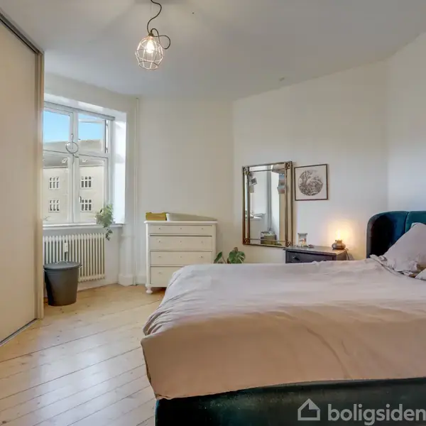 Soveværelse med en dobbeltseng dækket af puder. Spejlkommode og skænk står i baggrunden. Trægulv og vindue med udsigt. Loftlampe hænger centralt.