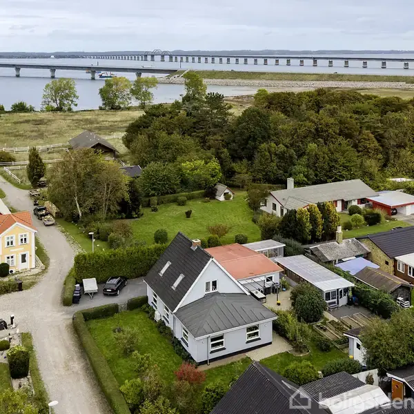 Luftfoto af et landsbyområde med flere huse omgivet af træer og veje, tæt ved en kystlinje. I baggrunden ses en lang bro, der strækker sig over vandet.