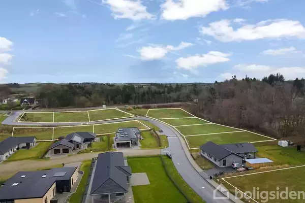 Flere moderne enfamiliehuse står langs en nyasfalteret vej i et parcelhuskvarter, omgivet af græsplæner og skovklædte områder under en blå himmel med skyer.