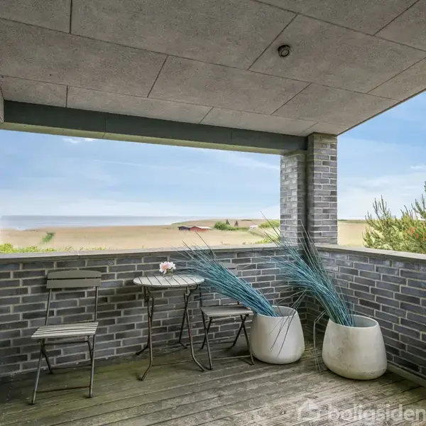 Balkon med grå murstensvægge, indeholder et lille bord med to stole og to hvide potter med planter. Udsigt til strand og hav med blå himmel i baggrunden.