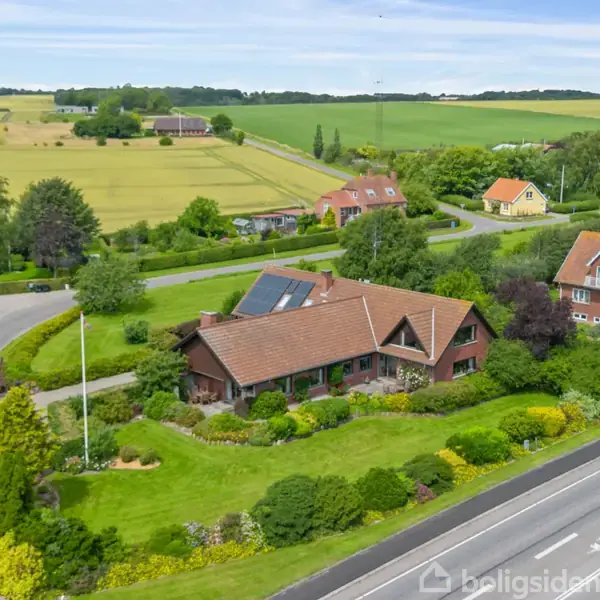 Et brunt hus med rødt tegltag ligger i en frodig have ved en vej. Omgivet af grønne marker, andre huse ses i baggrunden.