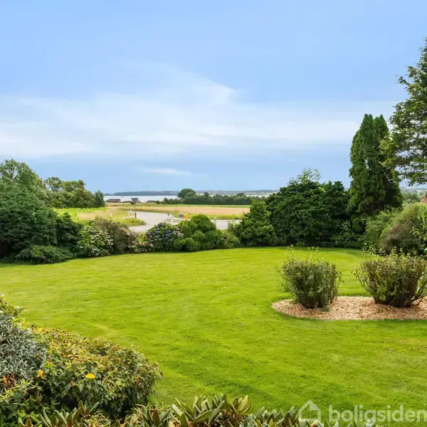 En grøn græsplæne strækker sig ud mod en række buske og træer. I baggrunden ses marker og en fjord under en blå himmel.