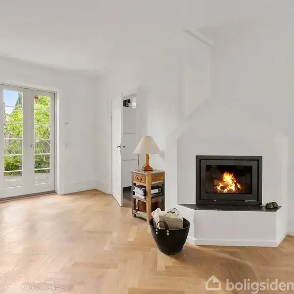 En pejs i et lyst rum brænder stille. Ved siden af står en lille reol med en lampe. Dobbeltfranske døre åbner ud mod en have.