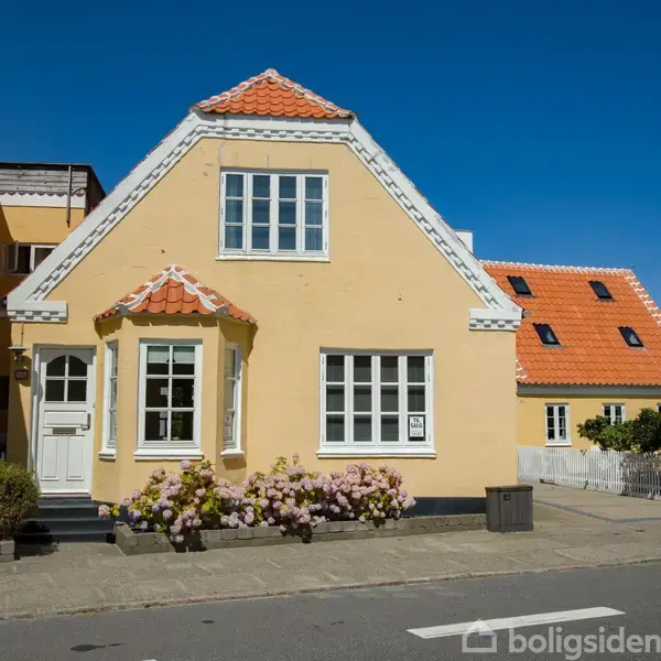 Gult hus med rødt tegltag, dør og to vinduer. Hus står ved vej med blomsterbede foran. Blau himmel uden skyer danner baggrund.