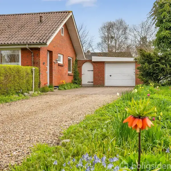 Rødt murstenshus med skråt tag står stille ved siden af en grusindkørsel. I forgrunden vokser farverige blomster blandt grønt græs. I baggrunden ses en garage og flere træer.