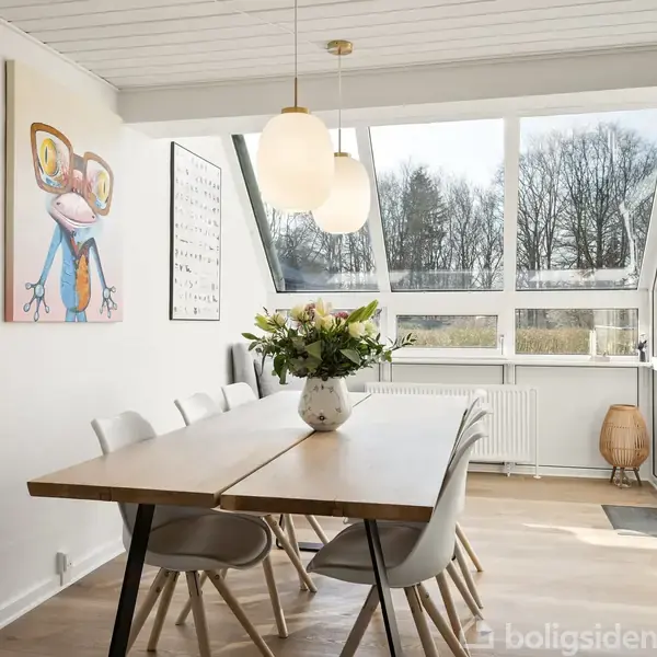 Et spisebord med stole står i et lyst rum. Ovenpå bordet er en vase med blomster. Væggene har kunstværker, og store vinduer giver udsigt til træer udenfor.