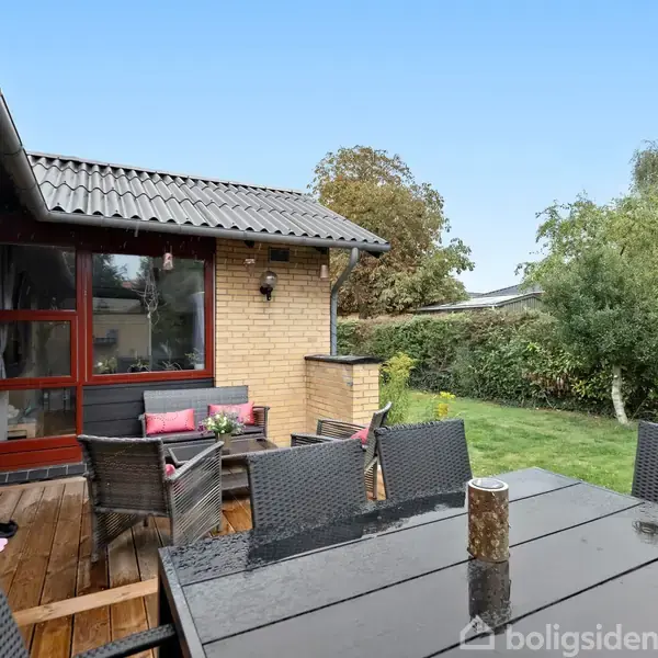 Sort havemøbelsæt med seks stole står på en træterrasse ved et gult murstenshus. Terrassebordet har en dekorativ genstand. Omgivet af græsplæne, buske og et træ.