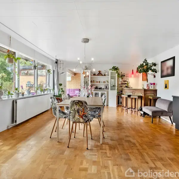 Spisestue med træbord og stole placeret midt i et lyst rum med trægulve. Vinduer langs venstre side giver udsigt til grøn have. Hylder og barområde bagest.