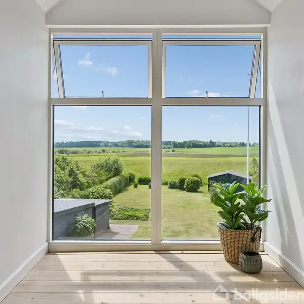 Et stort vindue viser udsigt over en grøn, frodig mark under en blå himmel. En plante står ved siden af vinduet på et lyst trægulv. Reol til venstre.