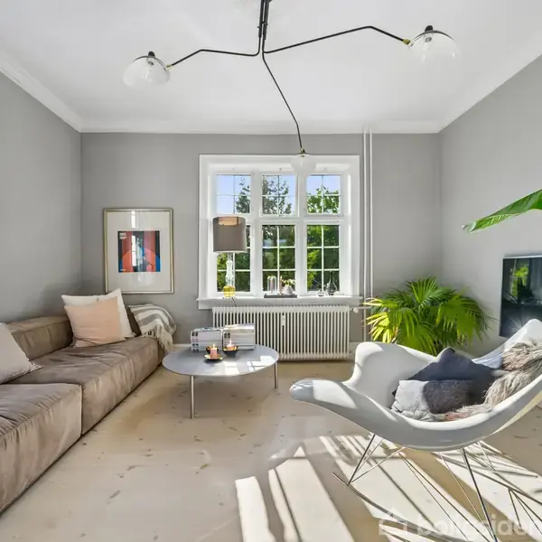 En grå sofa står i en stue med lyse vægge og gulv. En moderne lampe hænger fra loftet, mens et stort vindue lader sollys ind. Der er en grøn plante og et kunstbillede på væggen.