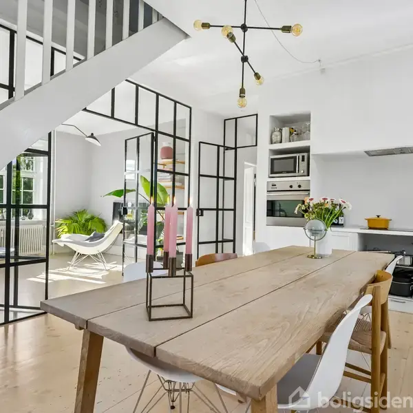 Spisebord af træ med stole omkring står i åbent køkken-alrum. Moderne lænestole og planter ses gennem glasvægge. Trappe på venstre side, enkelt belysning hænger fra loftet.