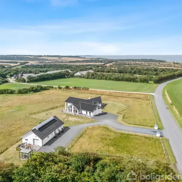 Et hvidt hus og en mindre bygning ligger ved en landevej omgivet af grønne marker. Landskabet er åbent med bølgende bakker i det fjerne under en blå himmel.
