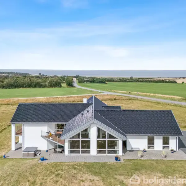 Et moderne hus med sort tag, store vinduer og en terrasse. Huset står i en landlig omgivelser med åbne marker og en vej i baggrunden under en klar blå himmel.