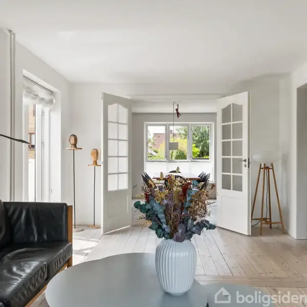 Sort lædersofa står foran lyse trædøre. Et rundt bord med en vase og tørrede blomster i forgrunden. Kunstværker hænger på de hvide vægge. Lysindfald fra vinduer.