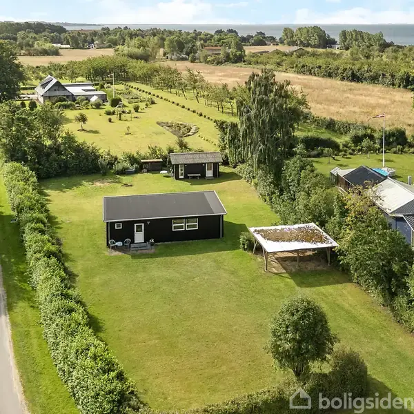 Et sort, rektangulært hus står midt i en grøn græsplæne med en hvid pergola ved siden af. Omkring ligger frodige marker og enkelte andre huse.