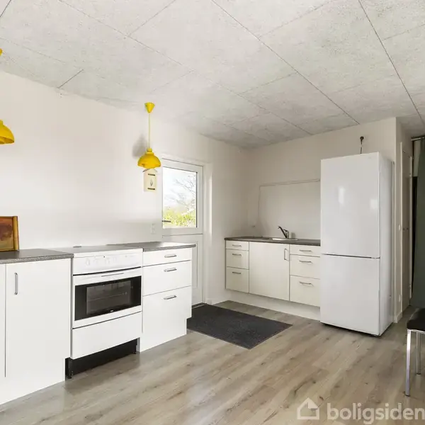 Et hvidt køkken med komfur og køleskab står i et rum med lyse træ-gulve. To gule lamper hænger fra loftet. En sort stol står i hjørnet.