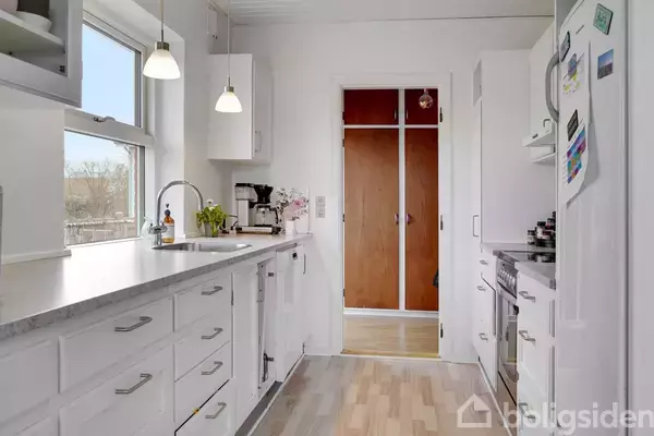 Et lyst køkken med hvide skabe og skuffer, står i et rummeligt rum. En kaffemaskine og planter pryder bordpladen under et vindue, mens ovnen er til højre.