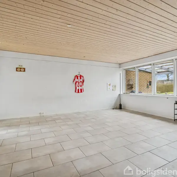 Et tomt rum med lyse fliser på gulvet og et lyst træloft. Der hænger en rød- og hvidstribet trøje på væggen. Store vinduer viser en have udenfor.