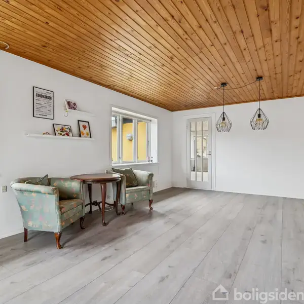 Et lyst rum med trægulv og træloft, indeholdende et bord og to lænestole ved vinduerne. To lamper hænger fra loftet, og på væggen ses hylder med billeder.