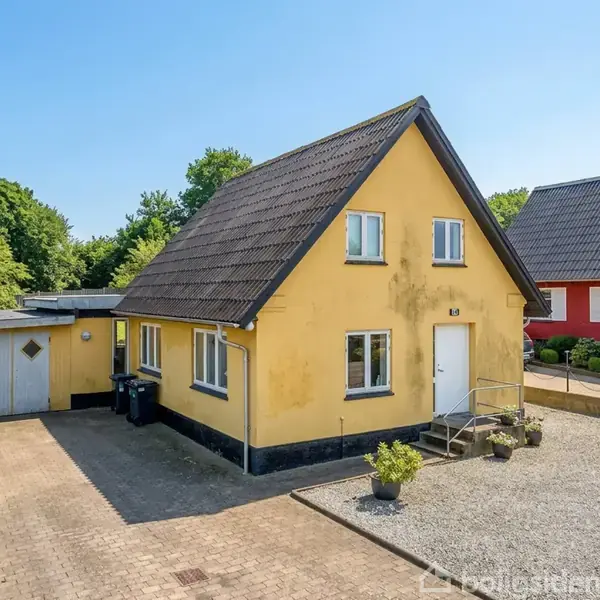 Et gult hus med spidst tag ligger i en boligkvarter. Huset har en hoveddør med trapper foran, og der er en dobbeltgarage til venstre. En grusbelagt forhave omkranser huset. Numret "83" er synligt på huset.