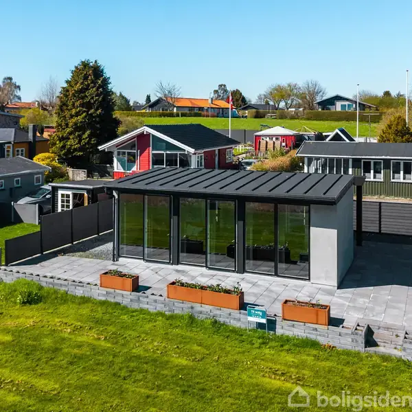 Et moderne, lille hus med store vinduer står på en terrasse omgivet af græsplæne og prydplanter i kasser. Haven ligger blandt andre lignende huse i et grønt område.