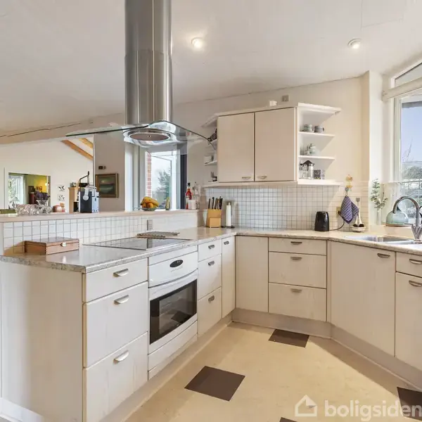 Et moderne, lyst køkken med hvide skabe og integreret ovn, står i et åbent rum. Omgivet af store vinduer med udsigt, forbindes det til en spisestue gennem en bred åbning.